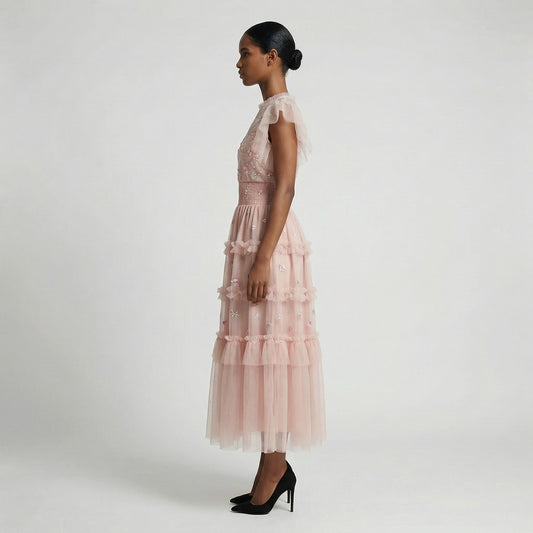 Full-length view of the Domenica pink mesh maxi dress featuring white and pink floral embroidery, a smocked waistline, and a tiered ruffled skirt laid out on a white faux fur rug.