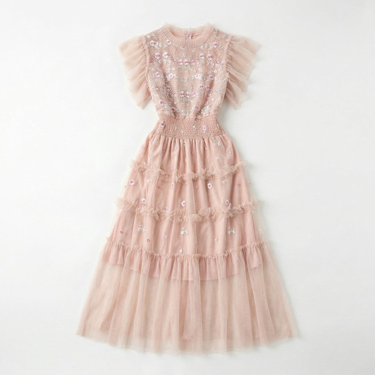 Full-length view of the Domenica pink mesh maxi dress featuring white and pink floral embroidery, a smocked waistline, and a tiered ruffled skirt laid out on a white faux fur rug.