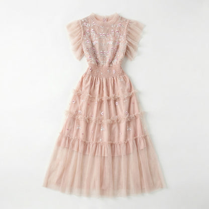 Full-length view of the Domenica pink mesh maxi dress featuring white and pink floral embroidery, a smocked waistline, and a tiered ruffled skirt laid out on a white faux fur rug.