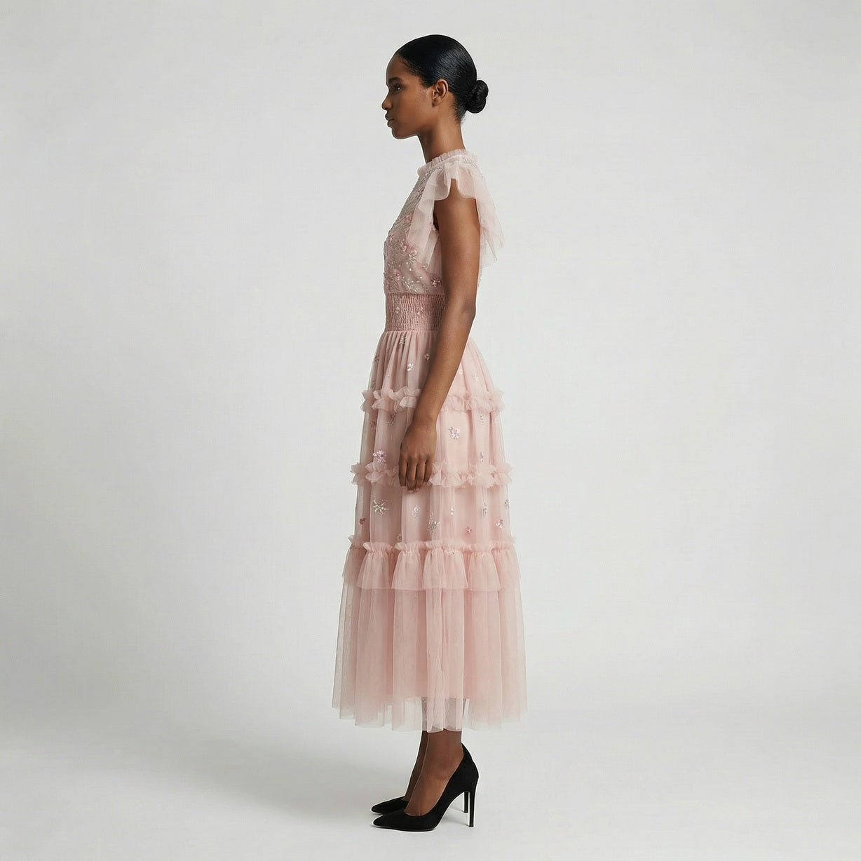 Full-length view of the Domenica pink mesh maxi dress featuring white and pink floral embroidery, a smocked waistline, and a tiered ruffled skirt laid out on a white faux fur rug.