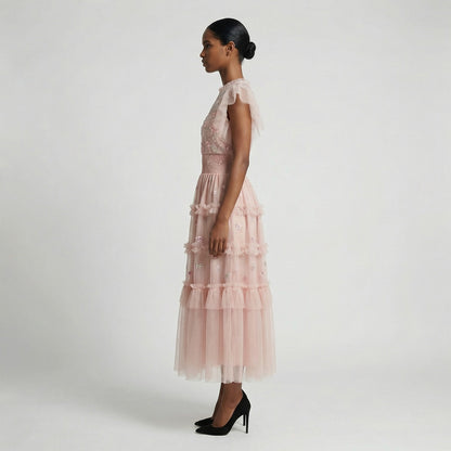 Full-length view of the Domenica pink mesh maxi dress featuring white and pink floral embroidery, a smocked waistline, and a tiered ruffled skirt laid out on a white faux fur rug.