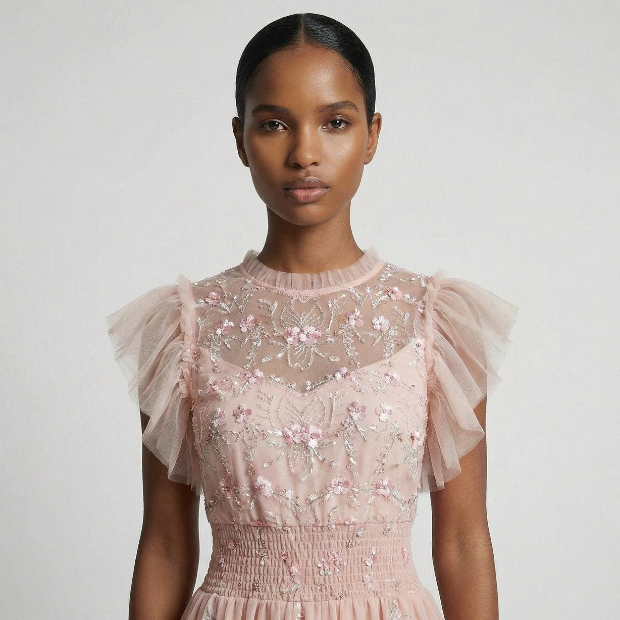 Full-length view of the Domenica pink mesh maxi dress featuring white and pink floral embroidery, a smocked waistline, and a tiered ruffled skirt laid out on a white faux fur rug.