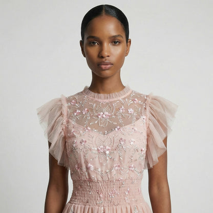 Full-length view of the Domenica pink mesh maxi dress featuring white and pink floral embroidery, a smocked waistline, and a tiered ruffled skirt laid out on a white faux fur rug.